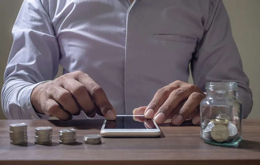 Un hombre que toca un teléfono móvil sobre un escritorio, con monedas en un tarro y apiladas sobre una mesa