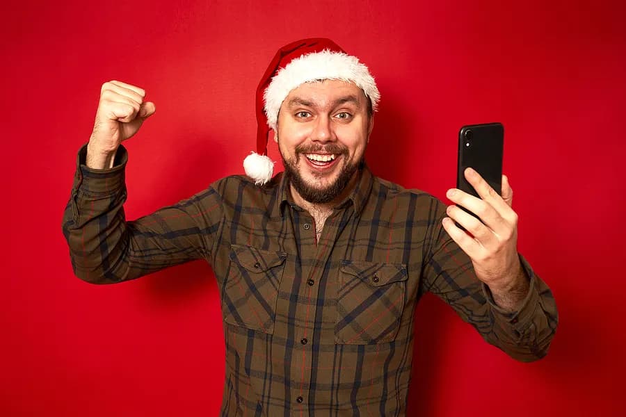 Hombre adulto con un gorro de Papá Noel, emocionado, sosteniendo un teléfono móvil