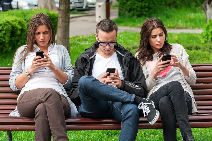 Tres personas sentadas en un banco del parque, entretenidas con sus teléfonos móviles.