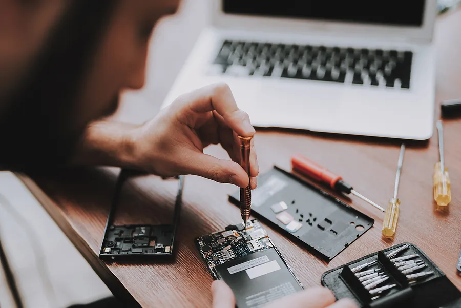 Un técnico utiliza un destornillador de precisión para reparar los componentes internos de un smartphone sobre un escritorio de madera, rodeado de pequeñas herramientas, piezas de recambio y un ordenador portátil abierto.