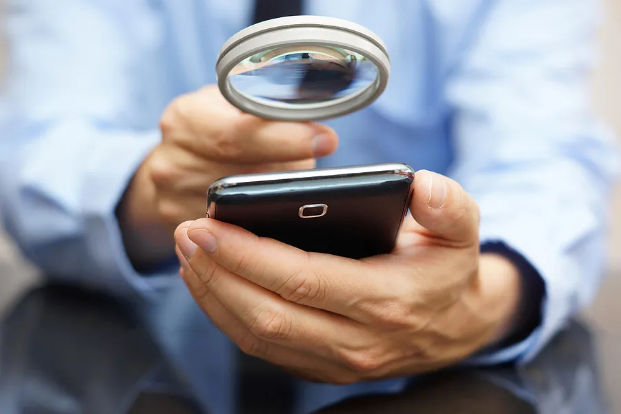 Una persona que examina detenidamente un smartphone con una lupa, comprobando la autenticidad del dispositivo o si presenta algún problema.