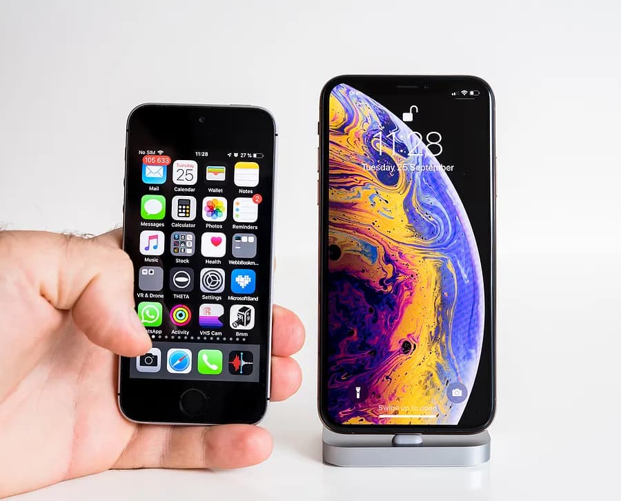 Una mano sostiene un iPhone pequeño y antiguo junto a un iPhone moderno y más grande colocado en un soporte, lo que permite apreciar las diferencias de tamaño y diseño entre un iPhone antiguo y un modelo más reciente.