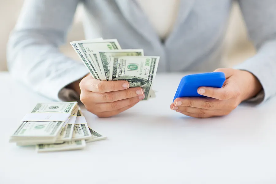 Persona que sostiene un smartphone en una mano y dinero en efectivo en la otra, lo que representa el valor de reventa del teléfono y la venta de un teléfono de segunda mano a cambio de dinero.